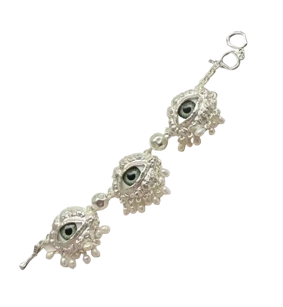 A silver-toned charm bracelet with three evil eye motifs and dangling pearl-like beads, displayed on a white stone slab against a dark background with a pink fabric corner.