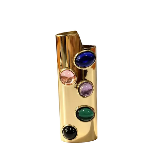 A polished gold cylindrical lighter or candle holder with five oval inset stones in blue, pink, purple, green, and black, standing on a white marble surface against a softly lit neutral background.