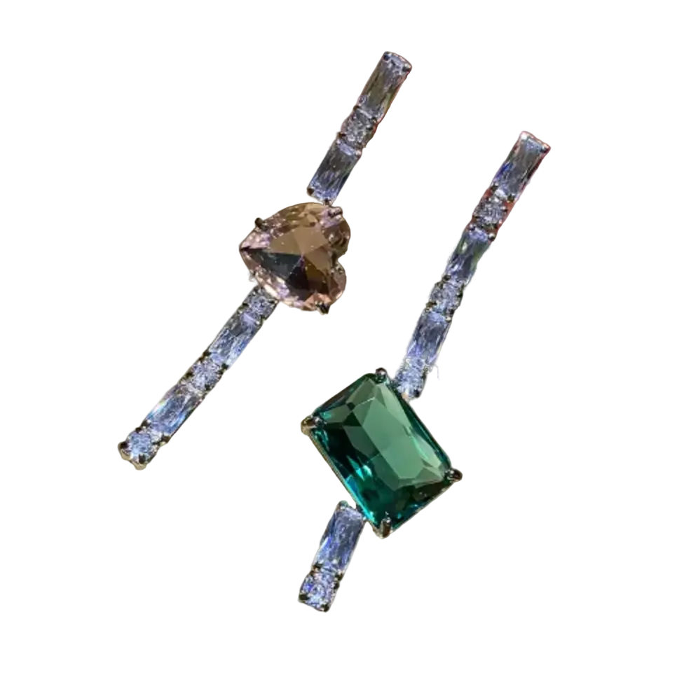 A close-up of a hand holding two drop earrings with silver-toned metal and clear stones. One earring features a heart-shaped pink gemstone, and the other features a rectangular emerald-cut green gemstone, both connected by vertical rows of clear baguette-cut stones.