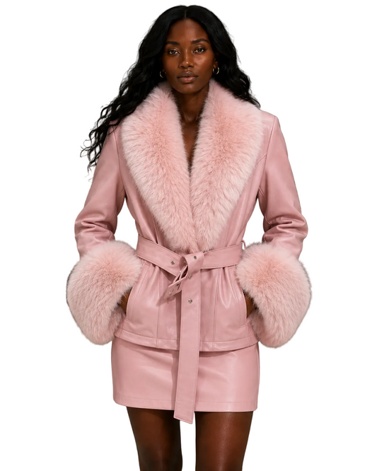 A model wearing a light pink faux-leather wrap coat with a large fluffy faux-fur collar and matching faux-fur cuffs, paired with a matching belt and mini skirt, set against a textured gray concrete wall.