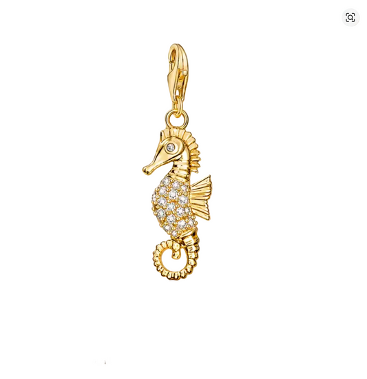 A gold seahorse-shaped charm or pendant with a small clear stone eye and a body paved with clear rhinestones, hanging against draped champagne satin fabric. The word THADDAEUS appears below, with smaller text 'CREATE YOUR OWN STYLE'.