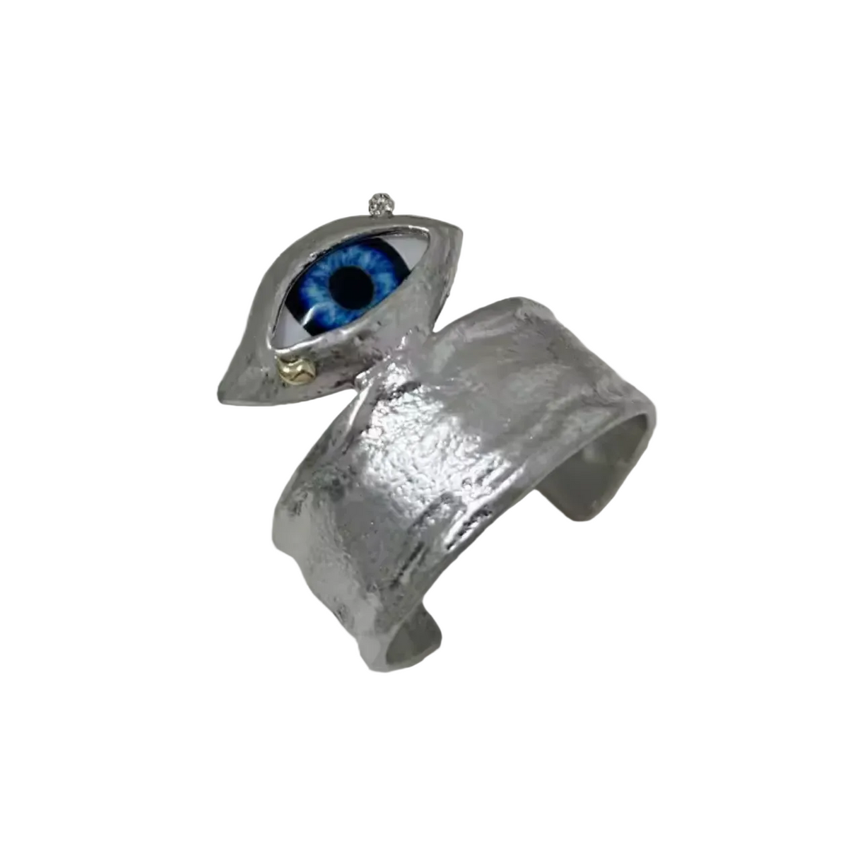 A textured silver cuff ring featuring a stylized eye charm with a vivid blue iris and a small gold accent, displayed on a light surface with a book in the background.