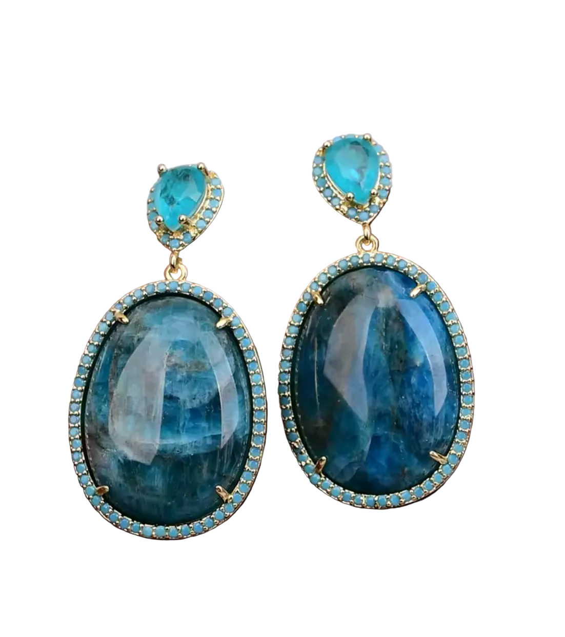 A pair of gold-tone drop earrings featuring large oval blue fluorite gemstones and smaller teardrop-shaped blue fluorite stones, both surrounded by small light blue cubic zirconia accents, displayed on a piece of brown wood against a soft pink background.